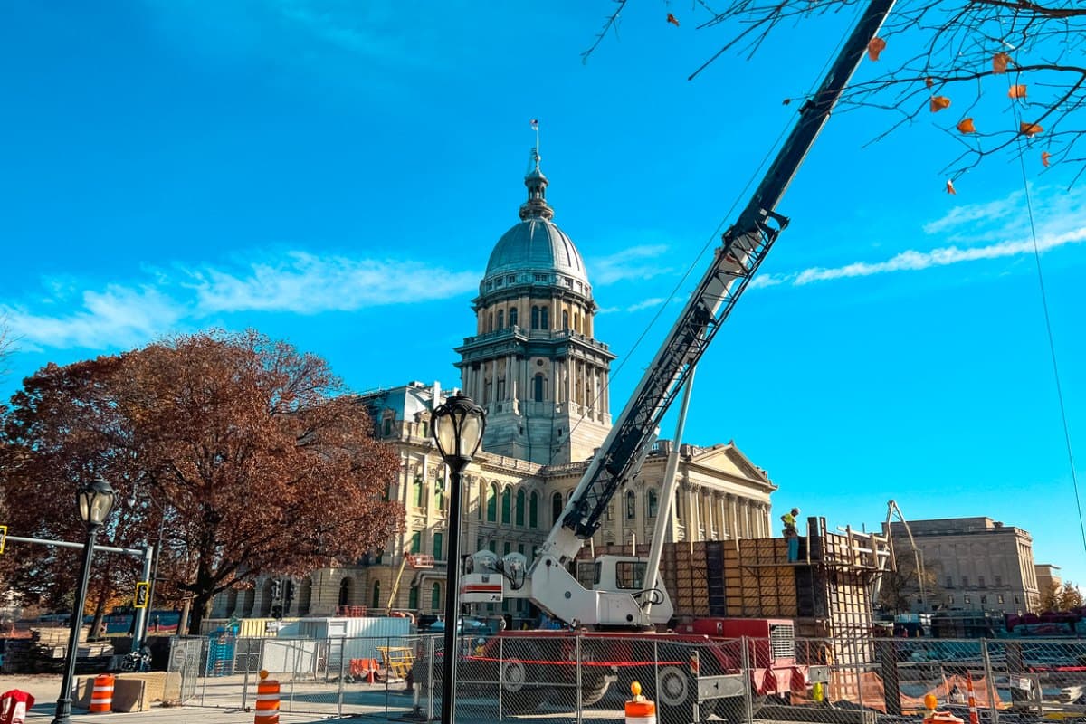 A mobile crane and fenced construction site in front of a domed state capitol building