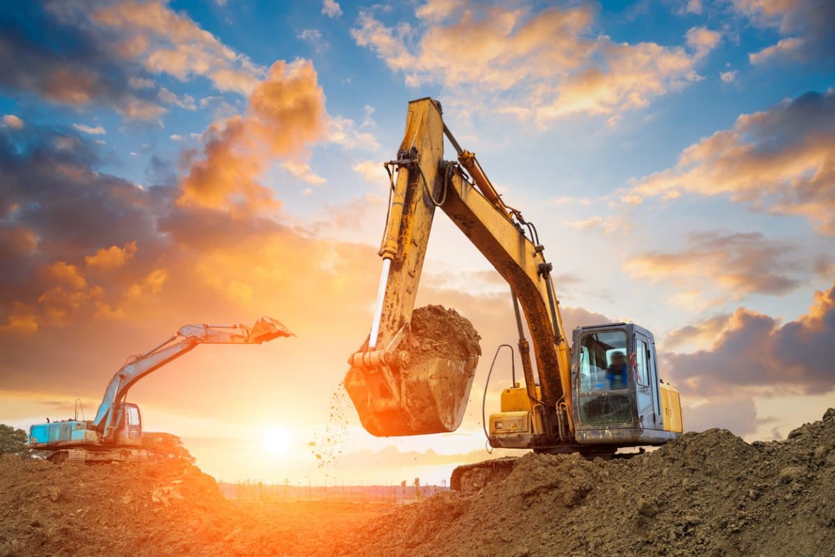 Two excavators moving earth on a site at sunset, one lifting a full bucket of soil