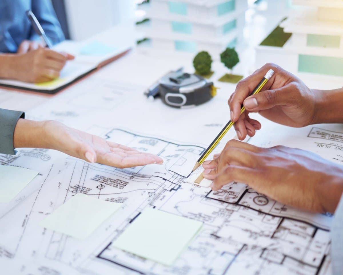 Two people reviewing a floor plan at a table, one marking the drawings with a pencil, with a tape measure and architectural models behind